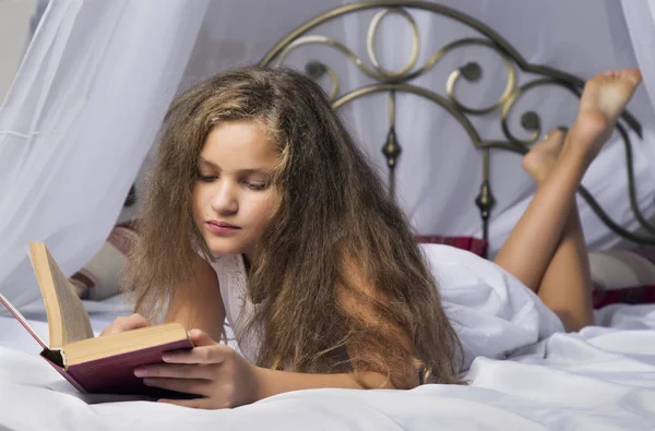 Nettes kleines Mädchen liest ein Buch und lächelt, während es auf einem Bett im Zimmer liegt — Stockfoto