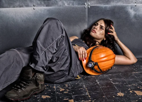 Beautiful tired sexy miner worker in orange helmet with flashlight lies on a floor on steel background