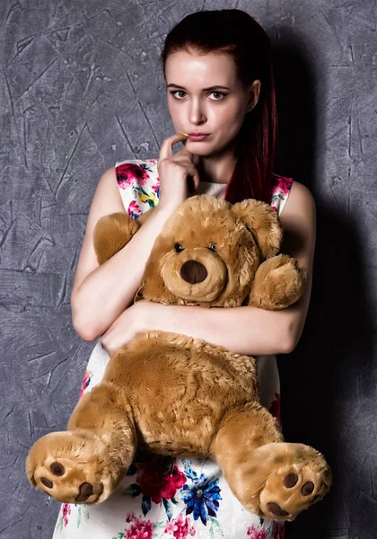 pensive or offended beautiful woman embraces a teddy bear on a gray background.