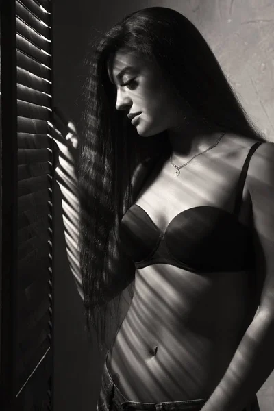 Hermosa mujer en ropa interior que se detiene cerca de la ventana y la persiana cerrada en un día soleado. Sombra rayada en un cuerpo — Foto de Stock