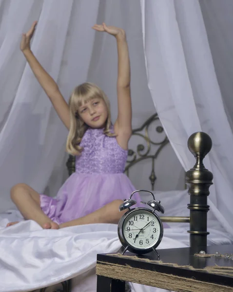 Alarm clock standing on bedside table. Wake up of an asleep young girl is stretching in bed in background — Stock Photo, Image