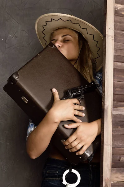 cowboy girl or pretty woman in stylish hat and blue plaid shirt holding gun and old suitcase