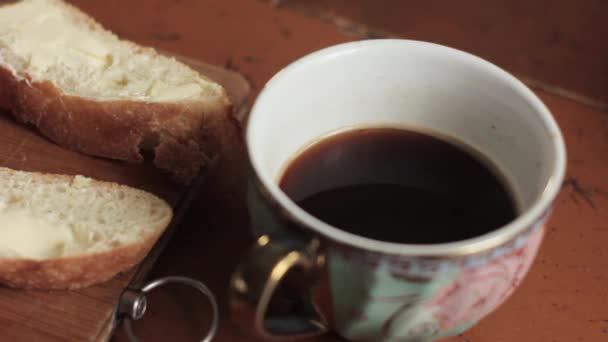 Burro su un pezzo di pane rurale e una tazza di caffè su un vecchio tavolo di legno. colazione — Video Stock