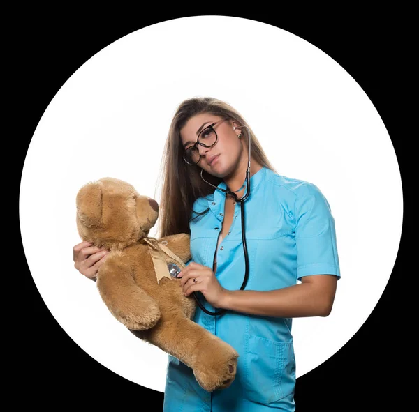 Sexy doctora con estetoscopio cura un oso de peluche sobre un círculo blanco y fondo negro —  Fotos de Stock