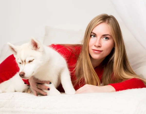 Schöne Frau im roten Kleid mit Husky-Welpen liegt auf einem weißen Bett — Stockfoto