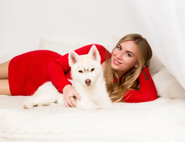 Hermosa mujer en un vestido rojo con husky cachorro se encuentra en una cama blanca —  Fotos de Stock