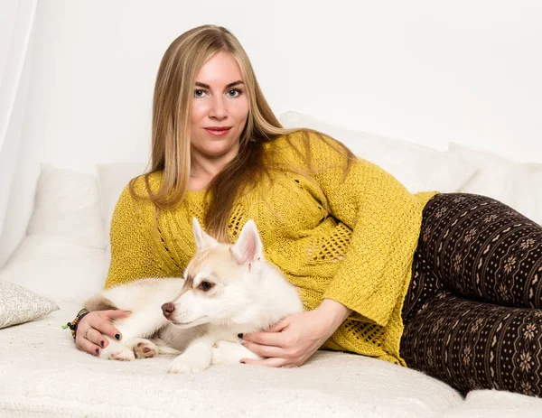 Blond kvinna i en pyjamas och ylle strumpor med husky valp ligger på en vit säng — Stockfoto