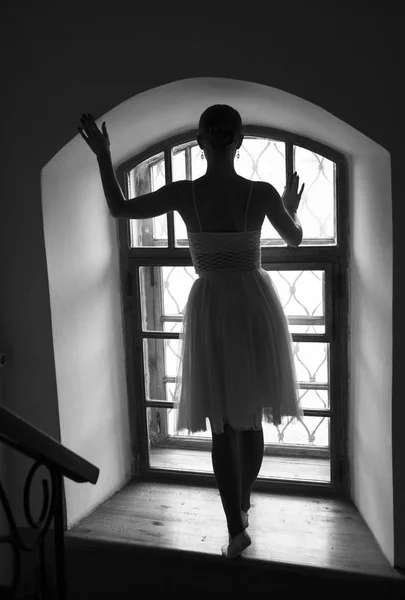 Bailarina jovem em vestido branco e sapatos de cetim balé posando em uma velha janela em um quarto escuro. preto e branco — Fotografia de Stock