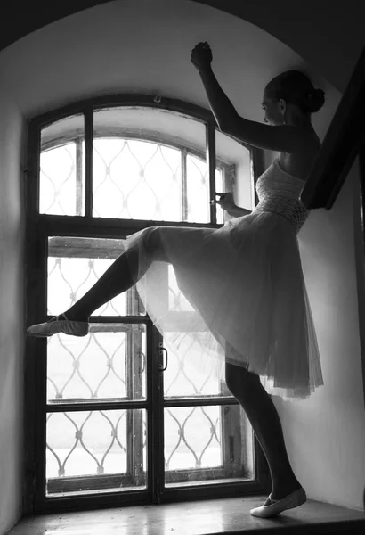 Junge Ballerina in weißem Kleid und Ballettschuhen aus Satin posiert an einem alten Fenster in einem dunklen Raum. schwarz-weiß — Stockfoto