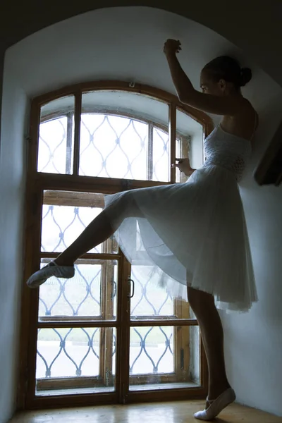Bailarina jovem em vestido branco e sapatos de cetim balé posando em uma velha janela em um quarto escuro — Fotografia de Stock