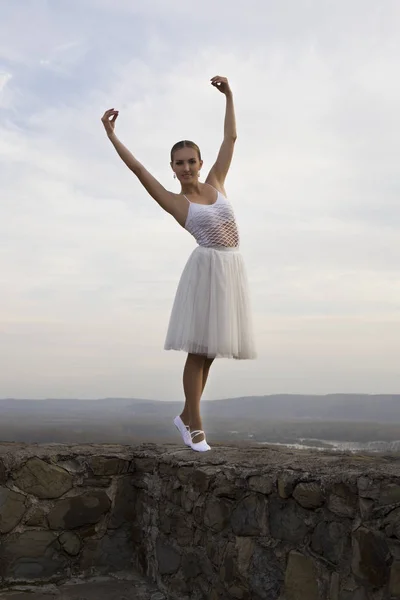 Jovem bailarina em vestido branco e sapatos de cetim balé posando na borda da antiga parede da fortaleza em um fundo de céu cinza — Fotografia de Stock