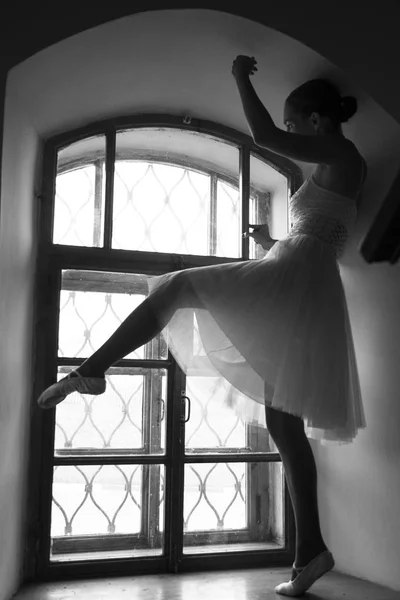 Jeune ballerine en robe blanche et chaussures de ballet en satin posant sur une vieille fenêtre dans une pièce sombre. noir et blanc — Photo