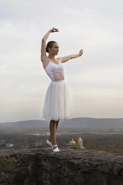 Jovem bailarina em vestido branco e sapatos de cetim balé posando na borda da antiga parede da fortaleza em um fundo de céu cinza — Fotografia de Stock