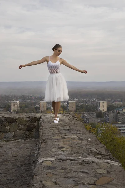 Jovem bailarina em vestido branco e sapatos de cetim balé posando na borda da antiga parede da fortaleza em um fundo de céu cinza — Fotografia de Stock