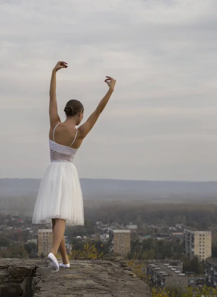 Jovem bailarina em vestido branco e sapatos de cetim balé posando na borda da antiga parede da fortaleza em um fundo de céu cinza — Fotografia de Stock