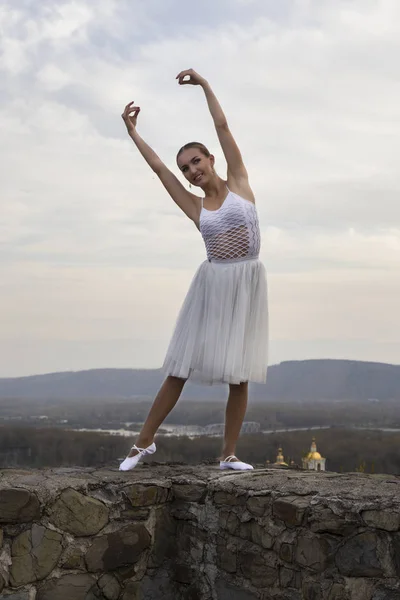 Jovem bailarina em vestido branco e sapatos de cetim balé posando na borda da antiga parede da fortaleza em um fundo de céu cinza — Fotografia de Stock