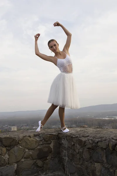 Jovem bailarina em vestido branco e sapatos de cetim balé posando na borda da antiga parede da fortaleza em um fundo de céu cinza — Fotografia de Stock