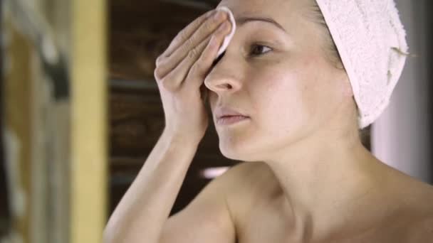 Mulher bonita em uma toalha tira a maquiagem na frente do espelho. Spa em casa — Vídeo de Stock
