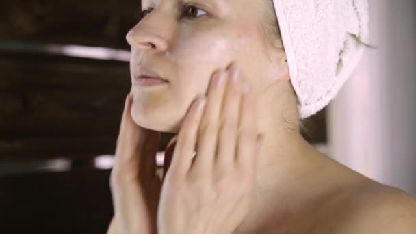 Mulher sã bonita em uma toalha pondo a nata que hidrata na cara. Cuidados da pele e casa Spa — Vídeo de Stock
