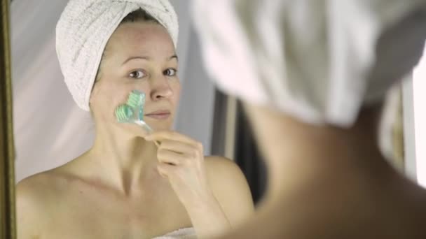 Красивая женщина в полотенце делает косметические процедуры в домашних условиях. массаж лица с массажером — стоковое видео