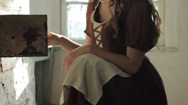 Vrouw in een rustieke jurk zat naast oude kachel in een verwoeste verlaten huis — Stockvideo