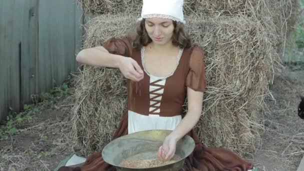 소박한 드레스 아름 다운 여자는 건초에 앉아서 곡물을 sifts — 비디오