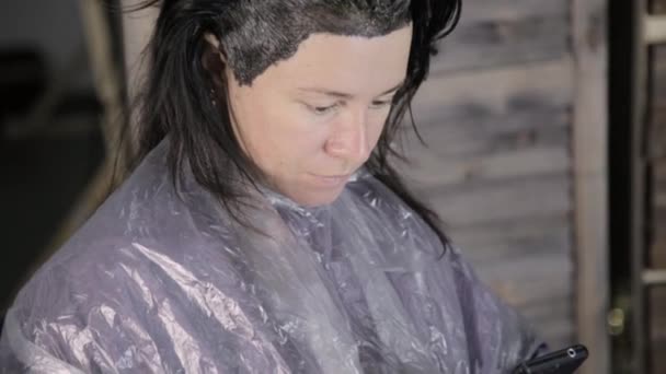 Hermosa mujer utiliza el teléfono inteligente en el salón de belleza, mientras que el trabajo de peluquería en su cabello — Vídeos de Stock