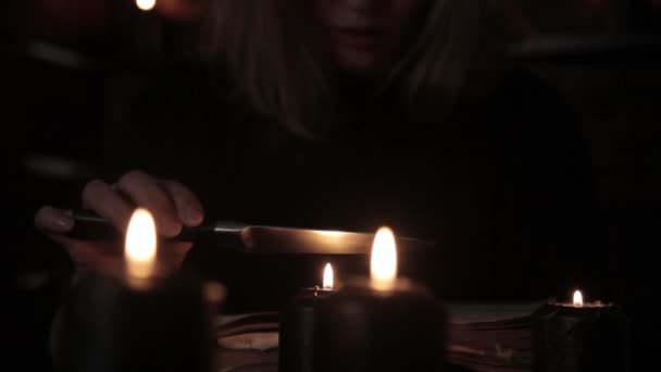 Bruxa detém uma faca e feitiço de leitura de livro velho mágico iluminado por velas pretas — Vídeo de Stock