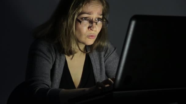Kvinna i glasögon använder dator i ett mörkt rum, ser på bildskärmen och börjar bli frustrerad — Stockvideo