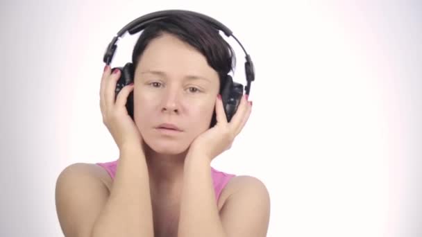 Brunette vrouw met grote koptelefoon muziek luisteren en dansen op een lichte achtergrond — Stockvideo