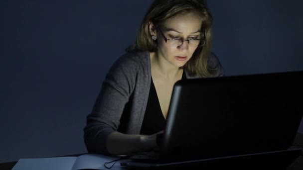 Mujer trabajadora que trabaja en el ordenador portátil y toma notas en el cuaderno en la habitación oscura — Vídeo de stock