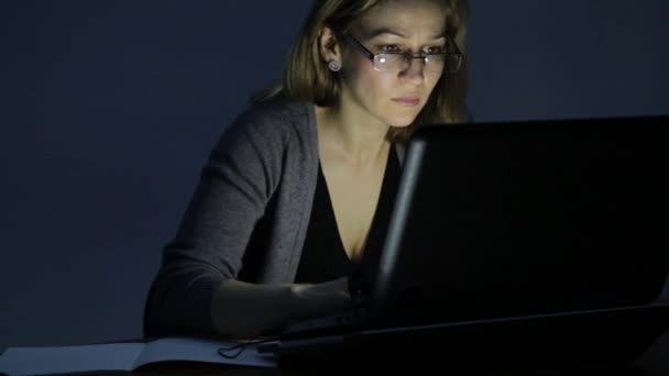 Fleißige Frau arbeitet am Laptop und macht sich Notizen in Notizbuch im dunklen Raum — Stockvideo