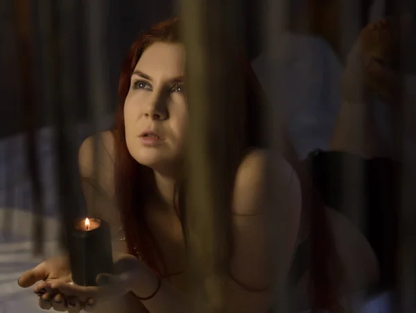 Sensual redhead woman lies on a bed and holding black burning candle — Stock Photo, Image