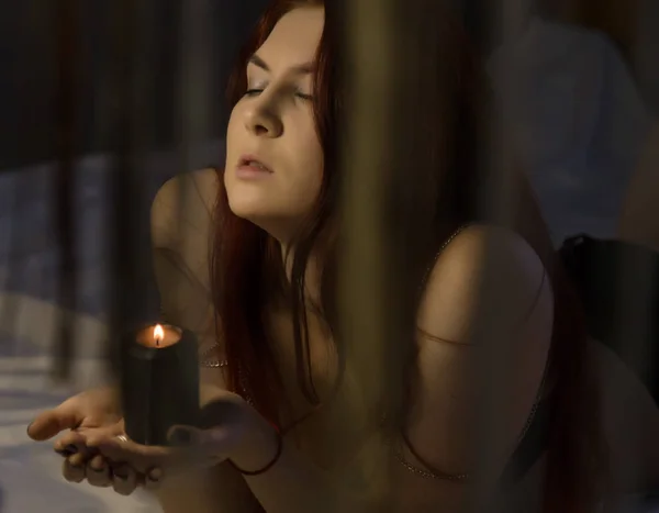 Sensual redhead woman lies on a bed and holding black burning candle — Stock Photo, Image