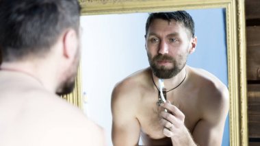 Sakallı adam banyoda aynanın önünde dişlerini fırçalıyor.