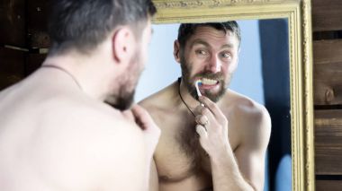 Sakallı adam banyoda aynanın önünde dişlerini fırçalıyor.