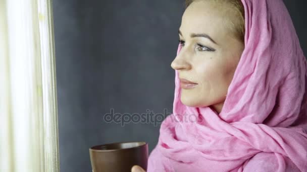 Красивая арабская мусульманка в розовом хиджабе пьет кофе перед окном — стоковое видео