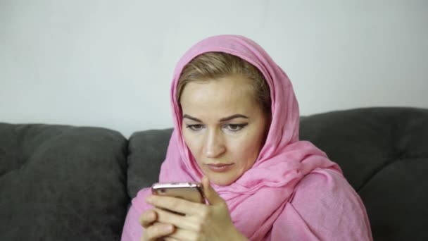 Beautiful arabian woman in pink hijab sitting on a sofa in a cafe and sends a text message on cell phone — Stock Video