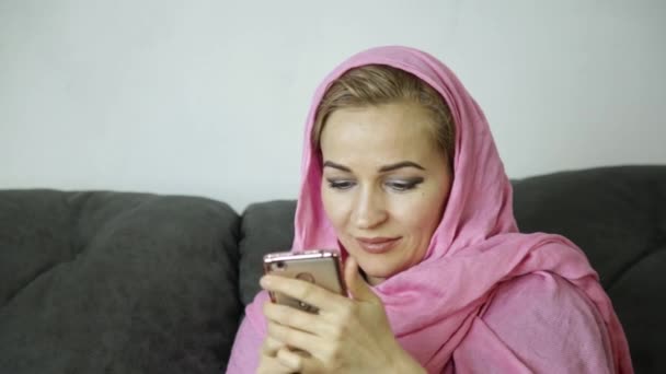 Молодая мусульманка в розовом хиджабе сидит на диване в кафе и отправляет смс-ку на сотовый телефон — стоковое видео