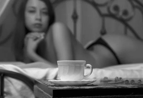 Sexy belle femme en sous-vêtements se détend sur un lit dans la chambre d'hôtel. gros plan d'une tasse de thé. noir et blanc — Photo