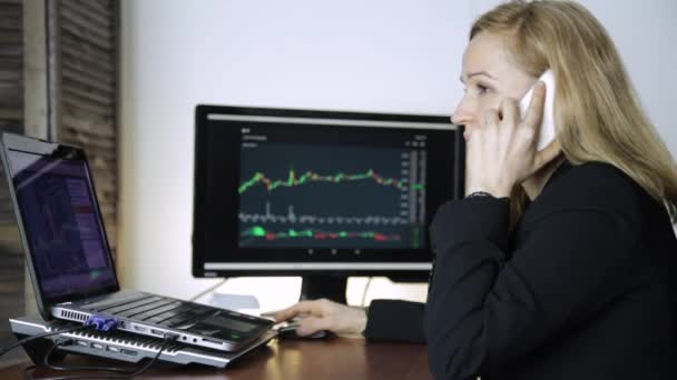 La dama de negocios aconseja al cliente en un teléfono y viendo el gráfico de cambio de divisas en el monitor de la computadora. Trabajar en una bolsa de valores criptográfica — Vídeos de Stock