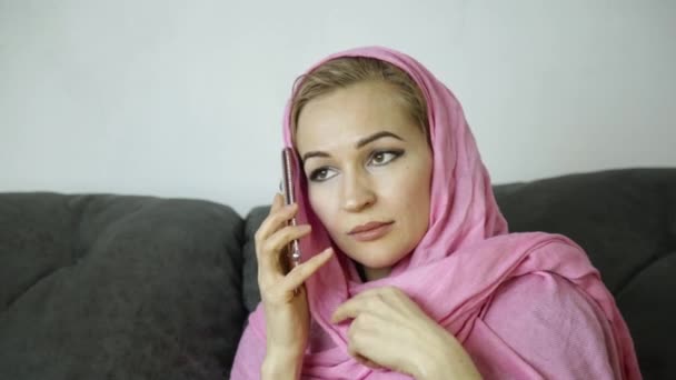 Mladá muslimka v růžové hidžáb sedí na pohovce v kavárně a mluví o mobilní telefon — Stock video