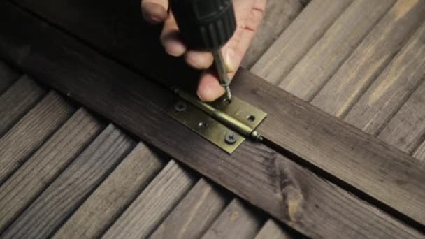 Человек прикрепляет дверную петлю к мебели с помощью электрической дрели — стоковое видео
