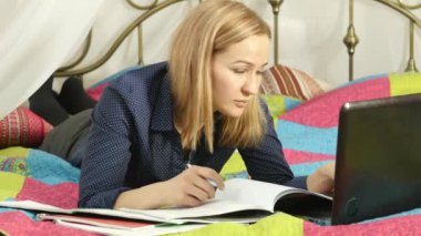 güzel bir kadın evinde on-line eğitim. Öğrenci çalışma kitabı ve bir dizüstü bilgisayar ile bir yatakta. 4k