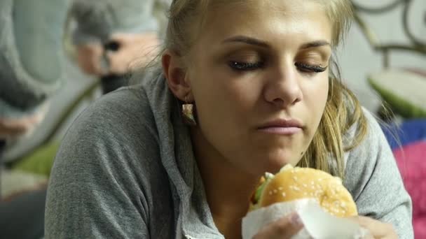 Nöjd kvinna äta snabbmat hamburgare på en säng i hennes rum. Mycket skräpmat. Slow motion — Stockvideo