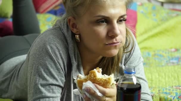 Rádi, že žena jíst fast food burger s šumivý nápoj na posteli ve svém pokoji. velmi nekvalitní potraviny. Zpomalený pohyb — Stock video