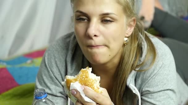 Довольная женщина ест гамбургер быстрого питания на кровати в своей комнате. очень вредная пища. Медленное движение — стоковое видео