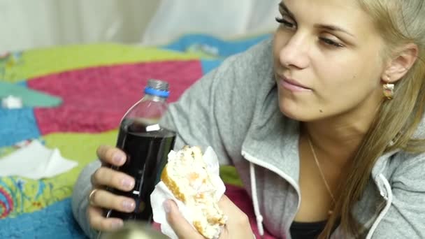 Задоволена жінка їсть бургер швидкого харчування з гарячим напоєм на ліжку у своїй кімнаті. дуже солодка їжа. Повільний рух — стокове відео