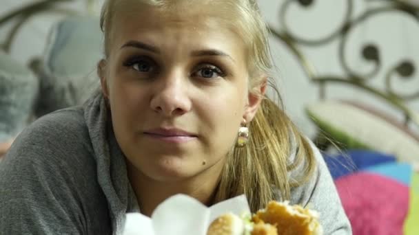 Довольная женщина ест гамбургер быстрого питания на кровати в своей комнате. очень вредная пища. Медленное движение — стоковое видео