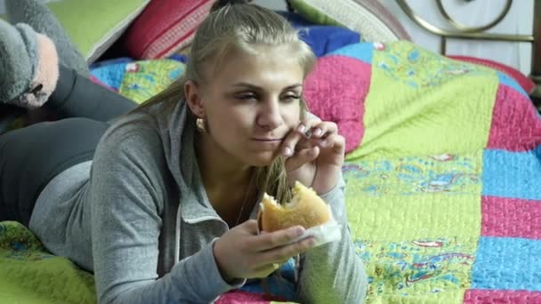 Mujer satisfecha comiendo hamburguesa de comida rápida en una cama en su habitación. muy comida chatarra. Movimiento lento — Vídeos de Stock